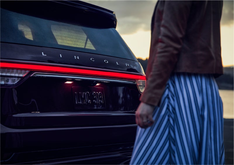 A person is shown near the rear of a 2023 Lincoln Aviator® SUV as the Lincoln Embrace illuminates the rear lights | Kindle Lincoln in Cape May Court House NJ