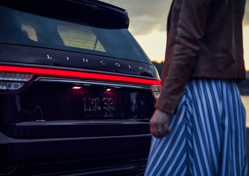 A person is shown near the rear of a 2024 Lincoln Aviator® SUV as the Lincoln Embrace illuminates the rear lights | Kindle Lincoln in Cape May Court House NJ